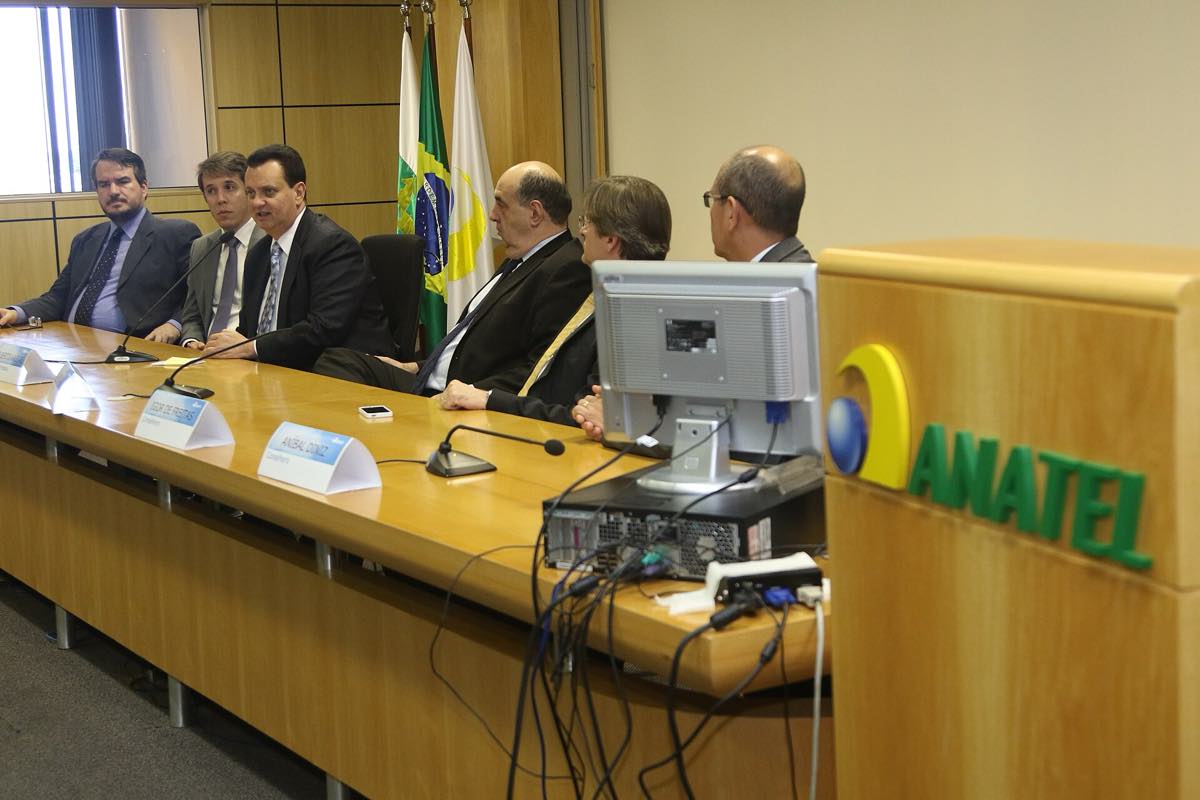 Mesa comprida de madeira clara com microfones e placas de identificação, seis pessoas de terno sentadas ao fundo, bandeiras do Brasil e um letreiro verde e amarelo com a palavra Anatel em primeiro plano.
