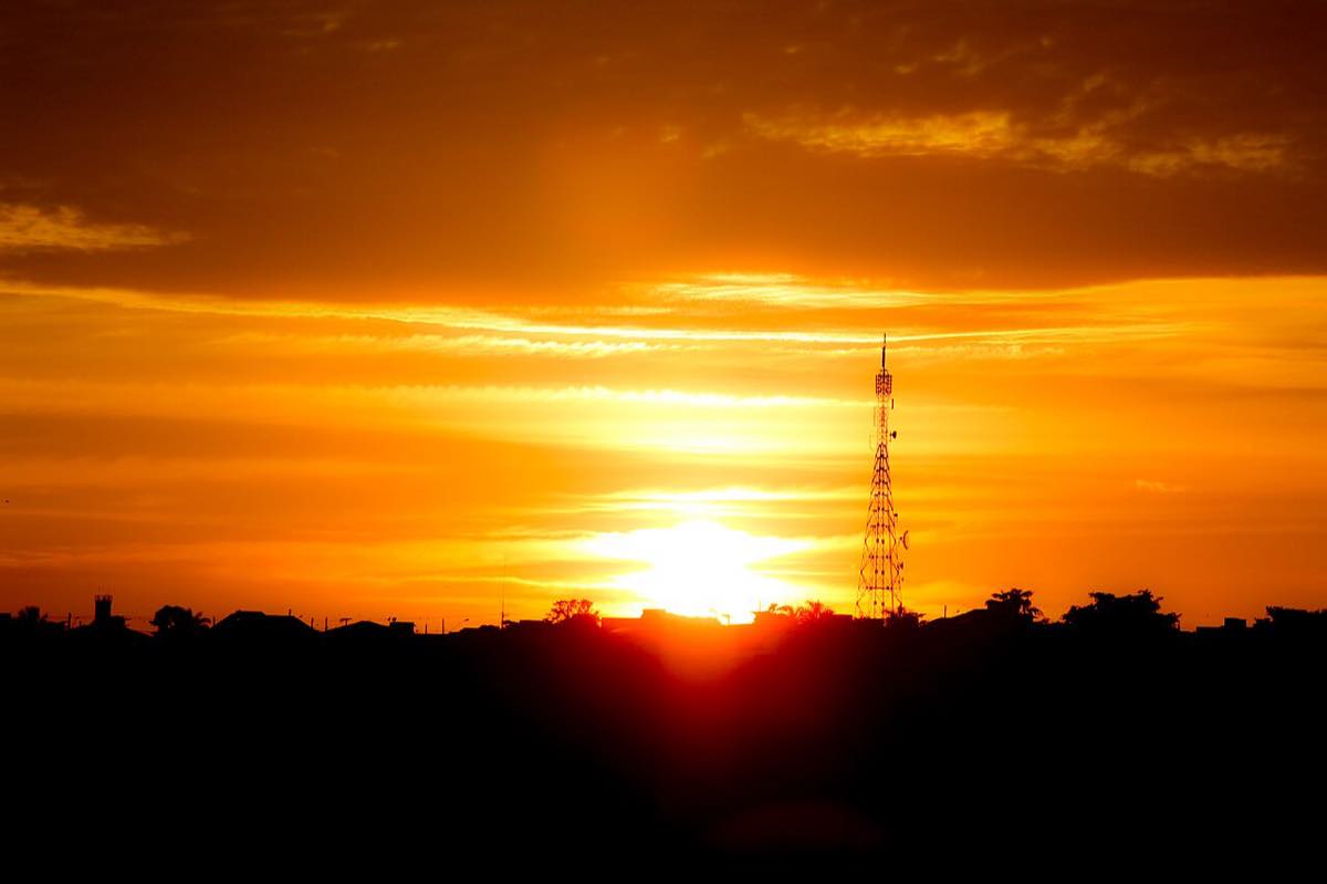 Amanhecer em Araxá, Minas Gerais: o sol nasce alaranjado atrás da silhueta escura da cidade, com uma torre de telecomunicações treliçada recortada contra o céu