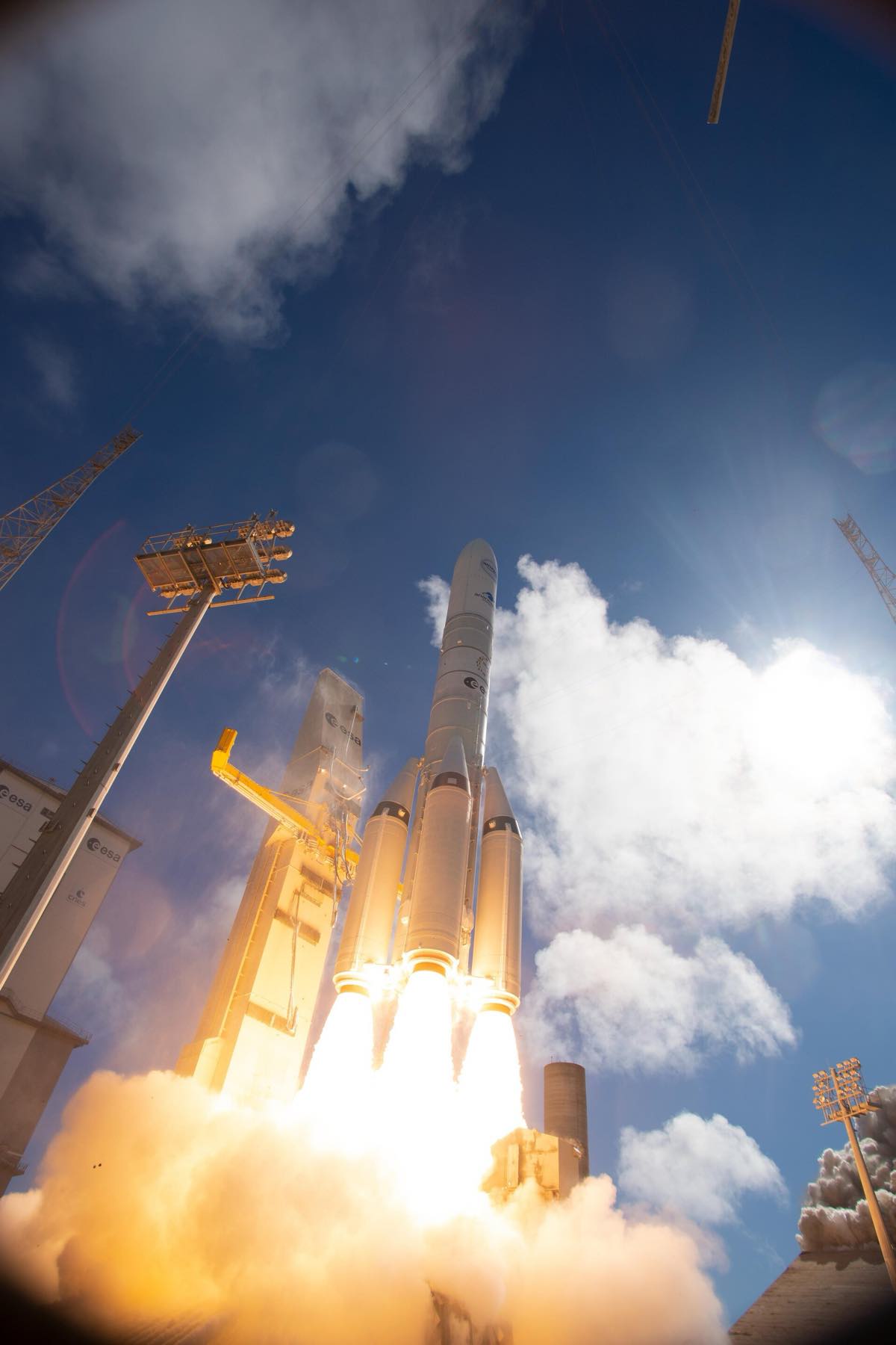 Foguete Ariane 6 fotografado de baixo no instante da decolagem, com chama alaranjada e nuvem branca de vapor tomando a base da plataforma, entre torres de iluminação do centro espacial.