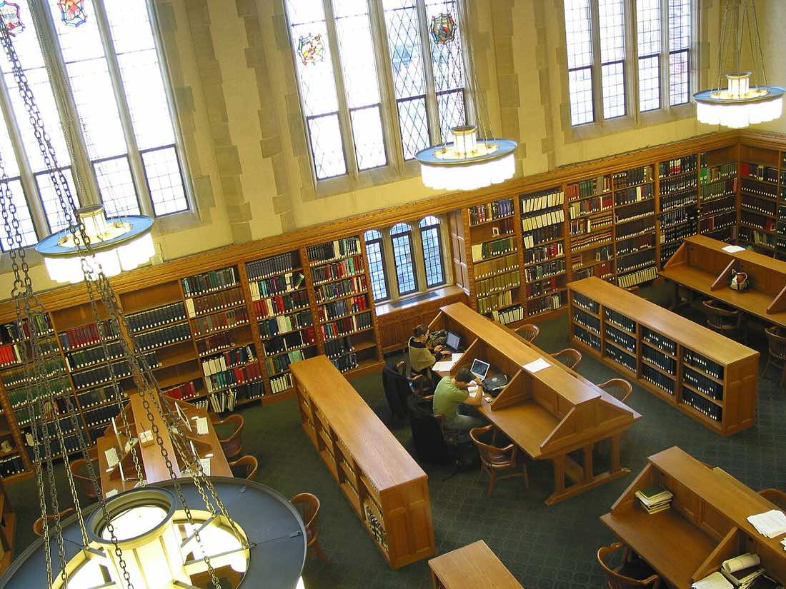 Sala de leitura de biblioteca jurídica com longas mesas de madeira, luminárias e estantes de livros.