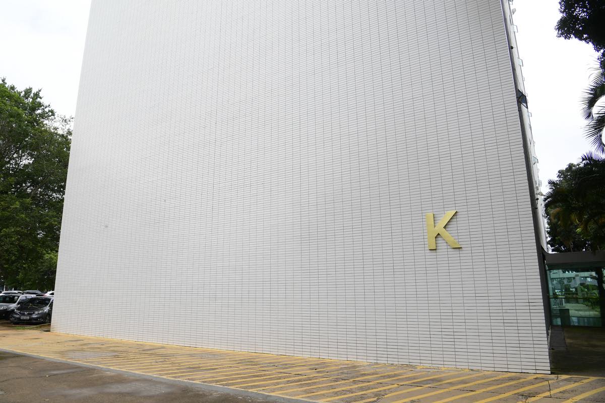 Fachada branca de azulejos do Bloco K da Esplanada dos Ministérios, em Brasília, com a letra K em dourado, árvores dos dois lados e faixas amarelas pintadas no chão