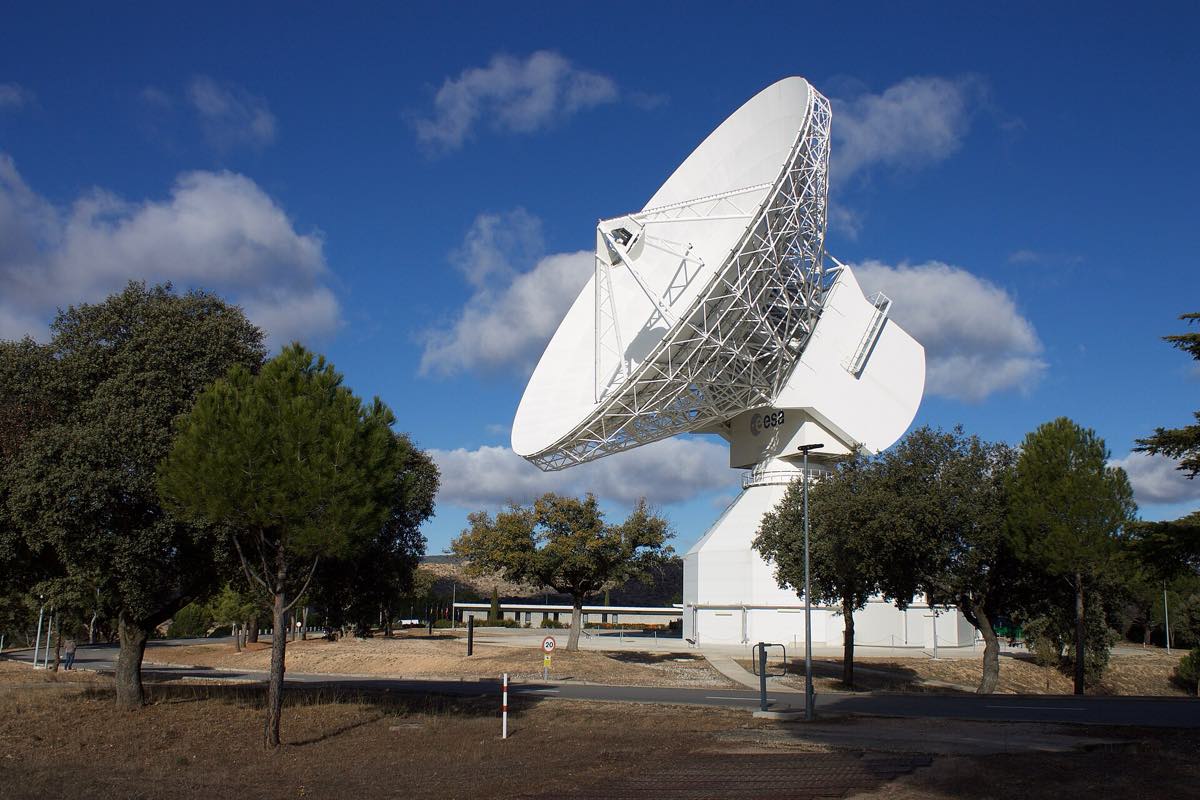 Grande antena parabólica branca de estação terrena da ESA, cercada de árvores, sob céu azul