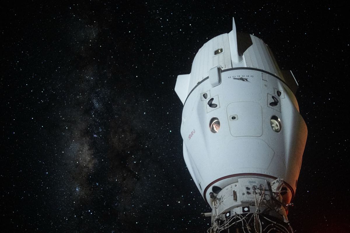 Cápsula Dragon da SpaceX, branca, acoplada à Estação Espacial Internacional, vista de baixo contra o fundo escuro do espaço com a Via Láctea e milhares de estrelas