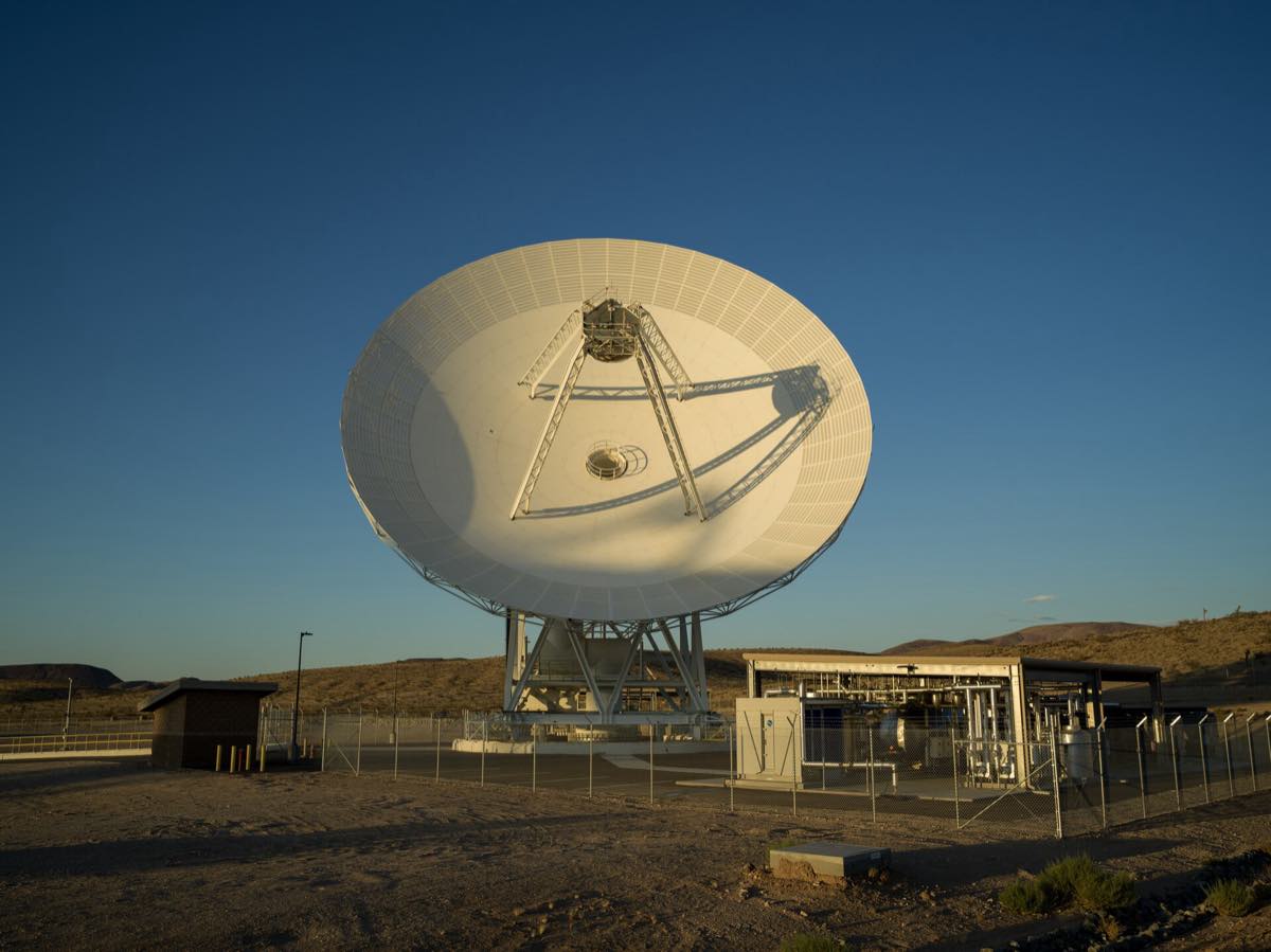 Antena parabólica branca de 34 metros do complexo de Goldstone, no deserto da Califórnia, com sombras longas ao entardecer.