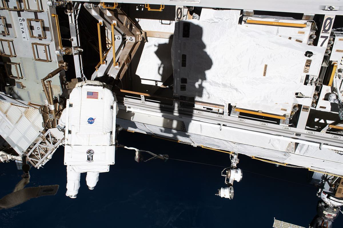 Astronauta de traje espacial branco, preso por cabo de segurança, flutua junto à estrutura externa da Estação Espacial Internacional; sobre a manta térmica branca ao lado projeta-se a sombra de outro astronauta, e ao fundo aparece o azul-escuro do oceano visto de órbita.