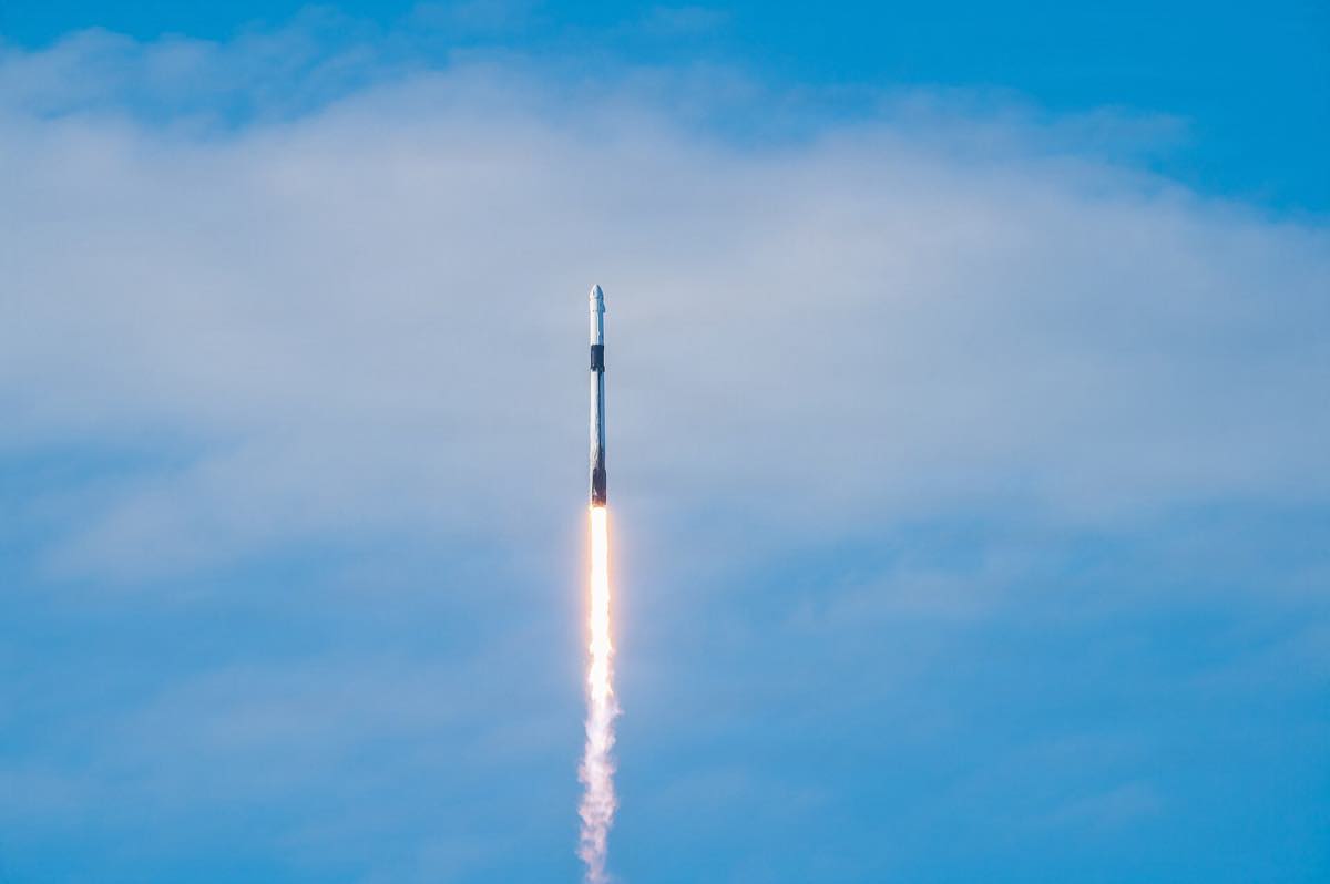 Um foguete Falcon 9 subindo contra um céu azul com nuvens, deixando um rastro de chama alaranjada