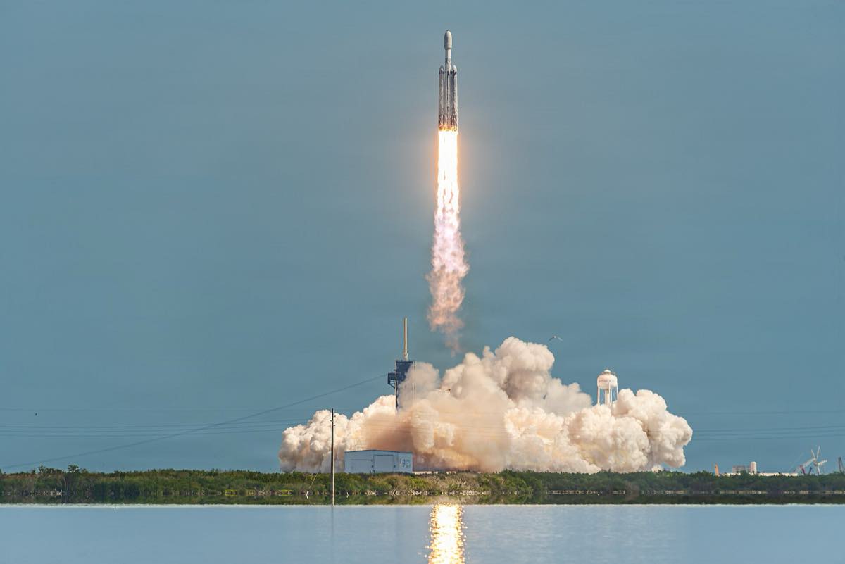 Foguete Falcon Heavy subindo com uma coluna de fogo e fumaça a partir da plataforma de lançamento, com o céu claro ao fundo.