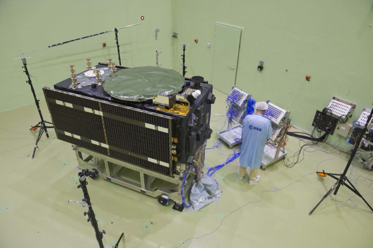 Um satélite Galileo em configuração de voo, com painéis solares dobrados, numa câmara de testes acústicos da ESA, com um técnico de jaleco azul ao lado