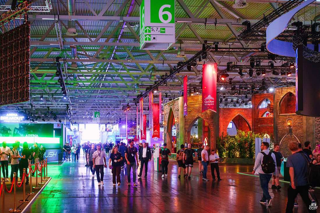 Corredor lotado de visitantes entre estandes iluminados na feira de games Gamescom 2024.
