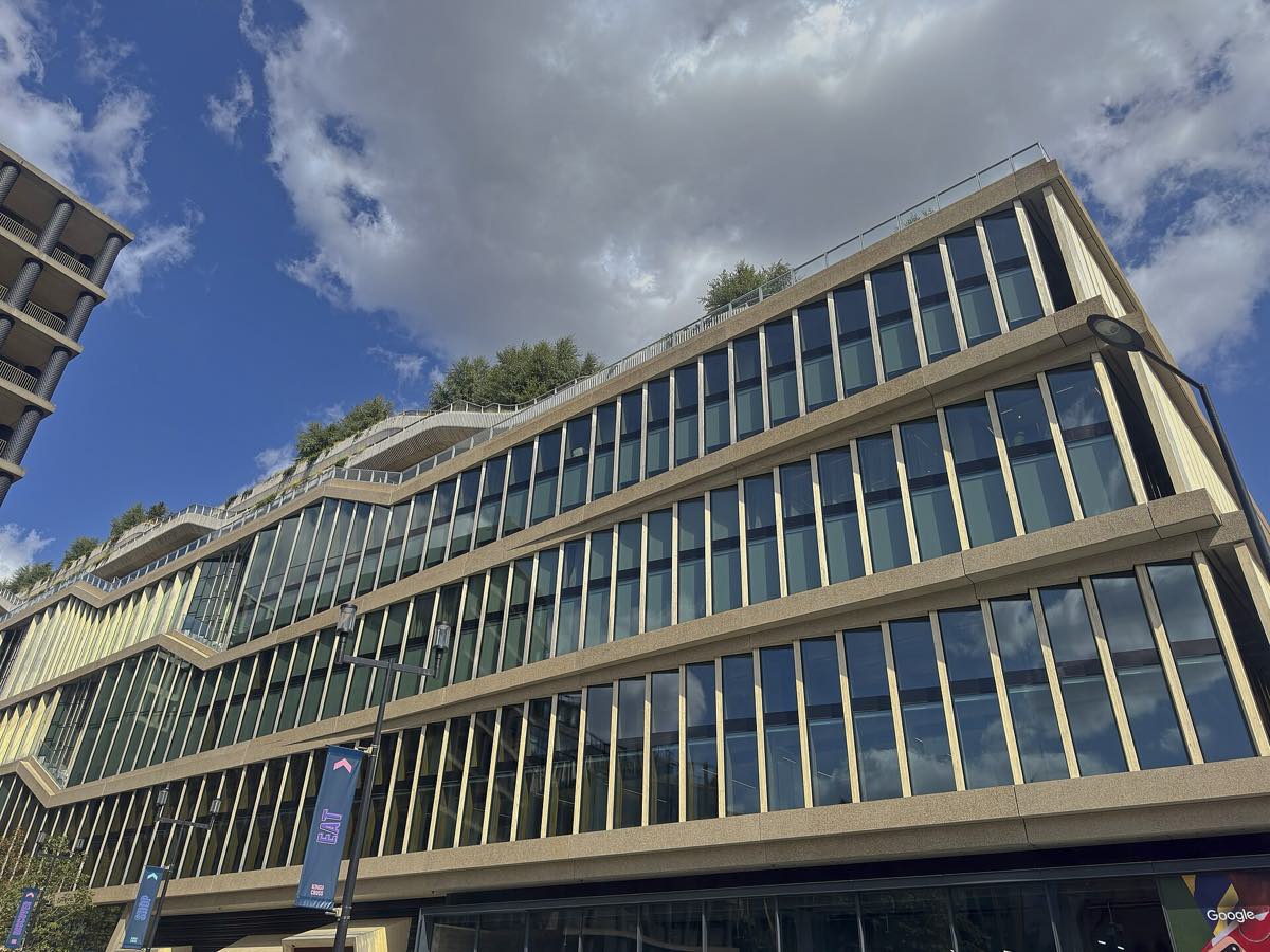Prédio comprido de vários andares, com fachada de vidro entre faixas claras, terraços com árvores no alto e o letreiro da Google no térreo, sob céu azul com nuvens