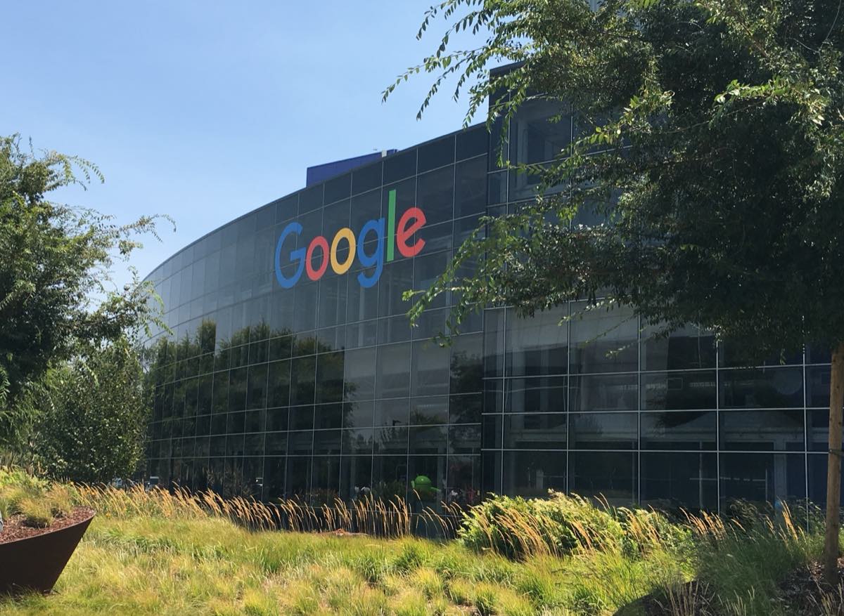 Fachada de um prédio do Googleplex, sede do Google em Mountain View, na Califórnia, com o logotipo colorido sobre a entrada e árvores ao redor