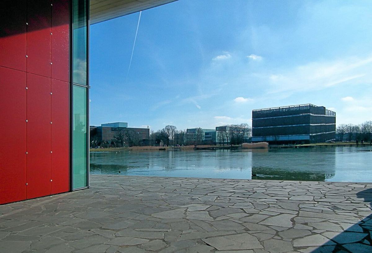 Vista do High Tech Campus Eindhoven a partir de um pátio coberto: uma parede de painéis vermelhos à esquerda, um lago à frente e prédios de escritórios envidraçados e um bloco escuro de fachada perfurada ao fundo, sob céu azul