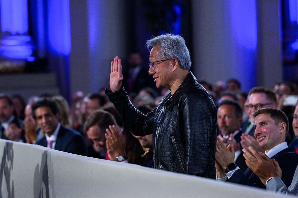 Jensen Huang, de jaqueta de couro, acena em pé durante evento sobre IA na Casa Branca, cercado por plateia que aplaude