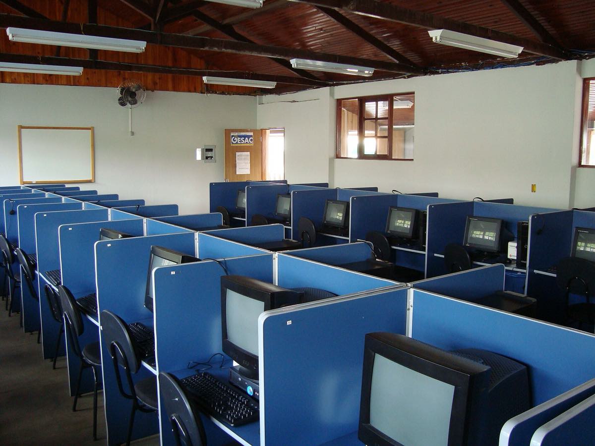 Sala com fileiras de computadores de mesa sobre bancadas, monitores desligados, em laboratório de informática no interior de Minas Gerais.