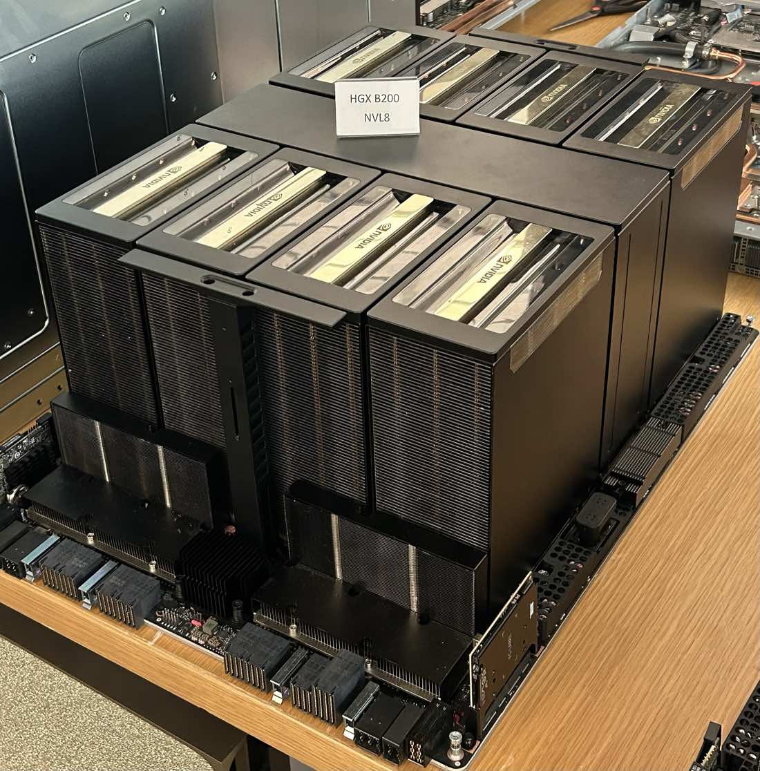 Placa de servidor com oito módulos de GPU pretos lado a lado, com dissipadores dourados no topo e uma plaquinha escrita HGX B200 NVL8, sobre uma bancada de madeira