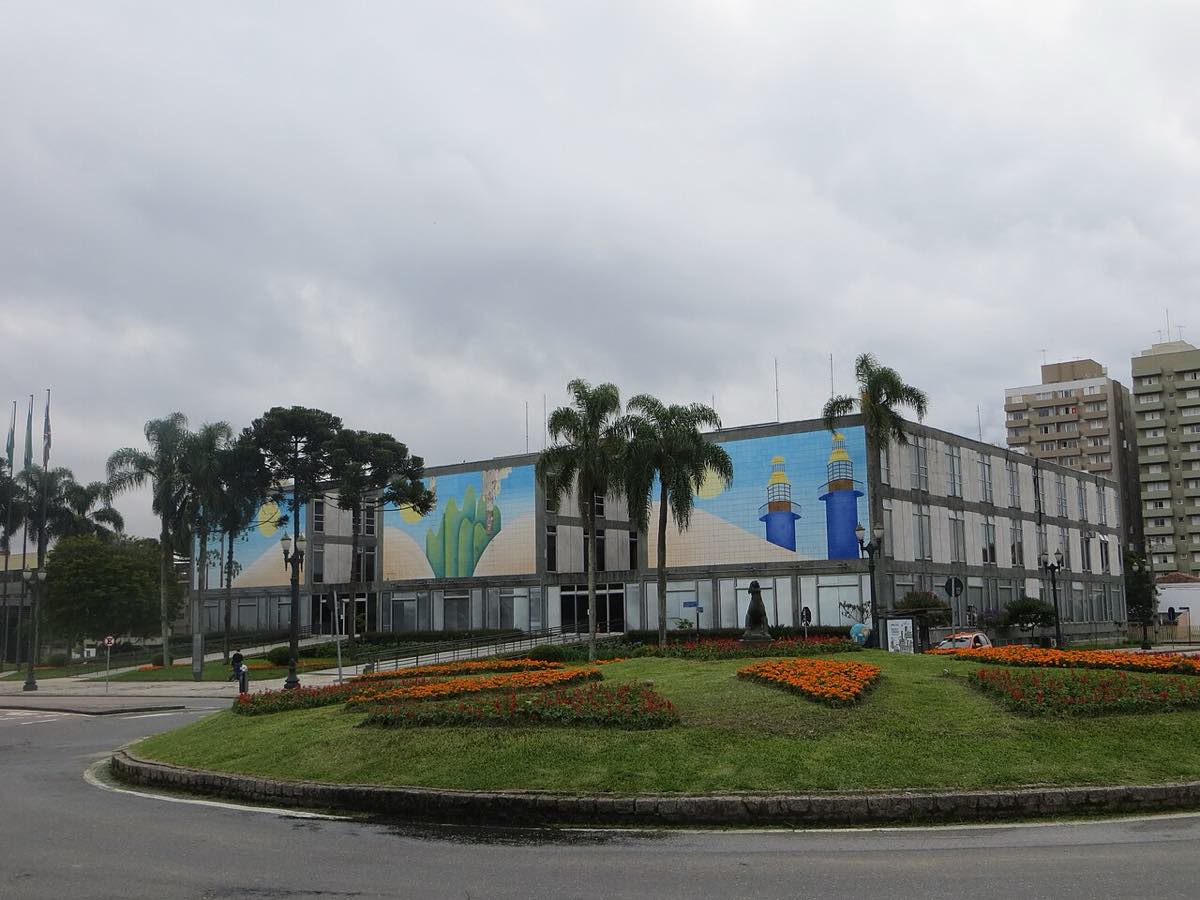 Fachada do Palácio 29 de Março, sede da Prefeitura de Curitiba, com painel colorido na fachada, palmeiras e canteiro de flores em primeiro plano