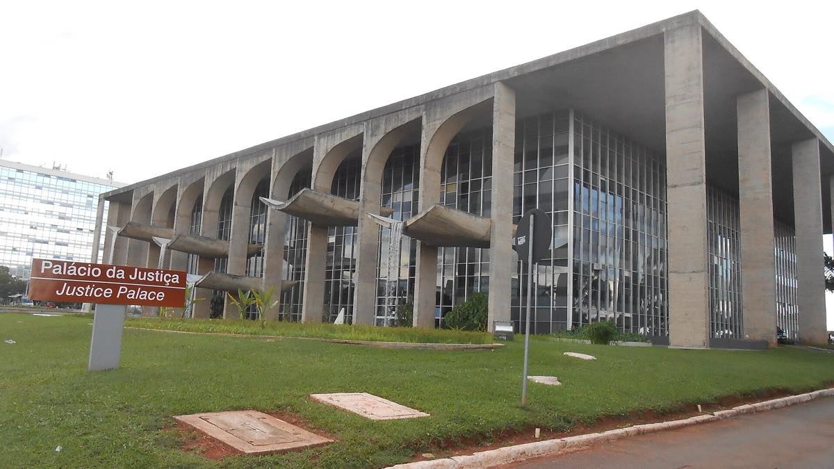Fachada do Palácio da Justiça em Brasília, com colunatas de concreto em arcos, calhas de água salientes e placa vermelha com os dizeres Palácio da Justiça na grama em primeiro plano.