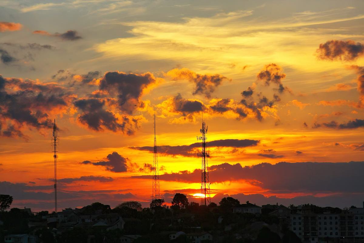 Pôr do sol alaranjado sobre Pato Branco, no Paraná, com três torres de telecomunicações recortadas em silhueta contra o céu e os telhados da cidade na parte de baixo do quadro.