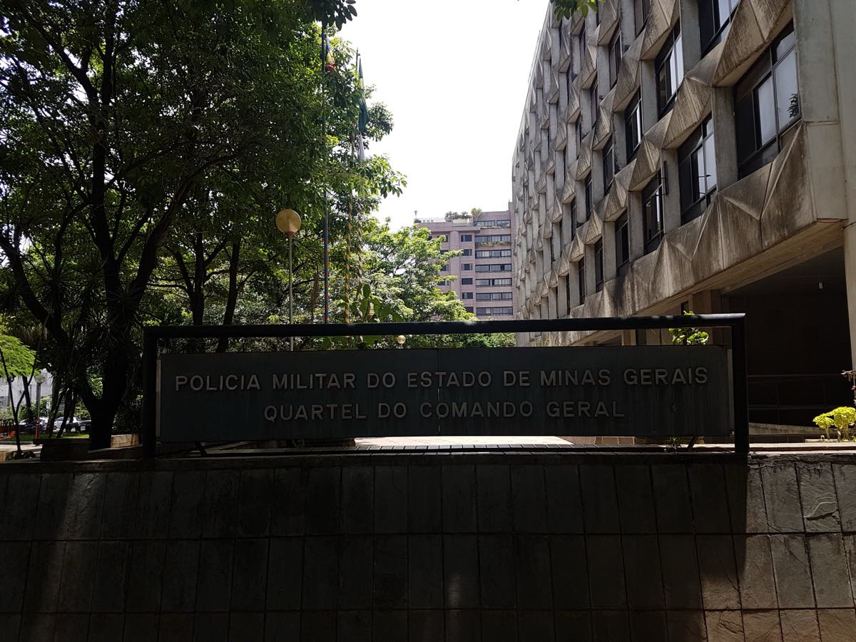 Placa de metal com os dizeres “Polícia Militar do Estado de Minas Gerais — Quartel do Comando Geral” diante de um prédio de concreto e árvores, em Belo Horizonte