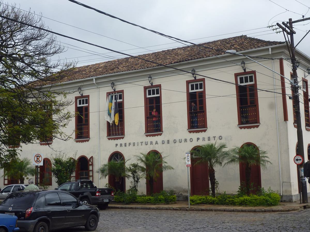 Fachada colonial branca com esquadrias escuras do prédio da Prefeitura de Ouro Preto, em Minas Gerais, sob céu claro.