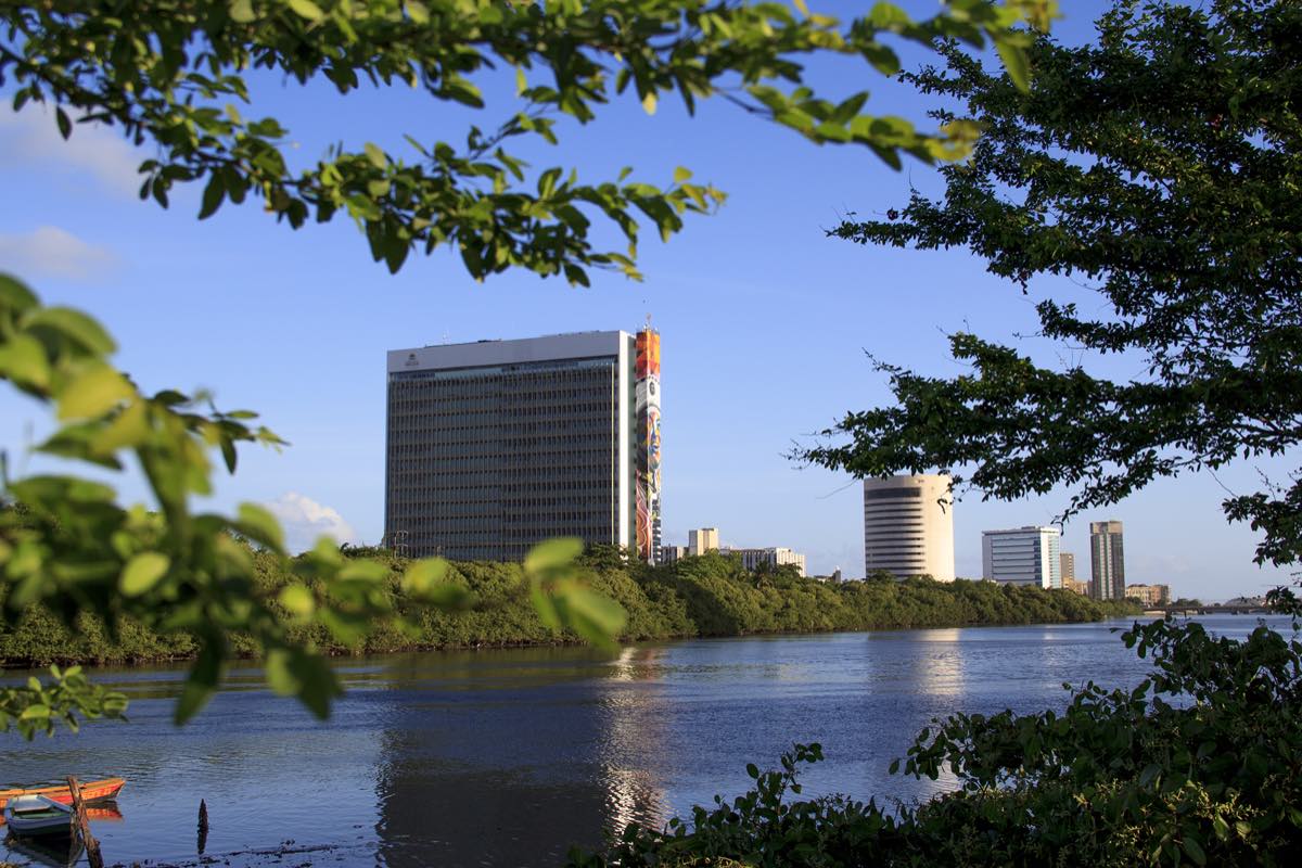 O edifício-sede da Prefeitura do Recife, uma torre clara de muitos andares com um mural colorido de um rosto ocupando toda a lateral, visto do outro lado do rio Capibaribe, com mangue na margem e um barco pequeno em primeiro plano.