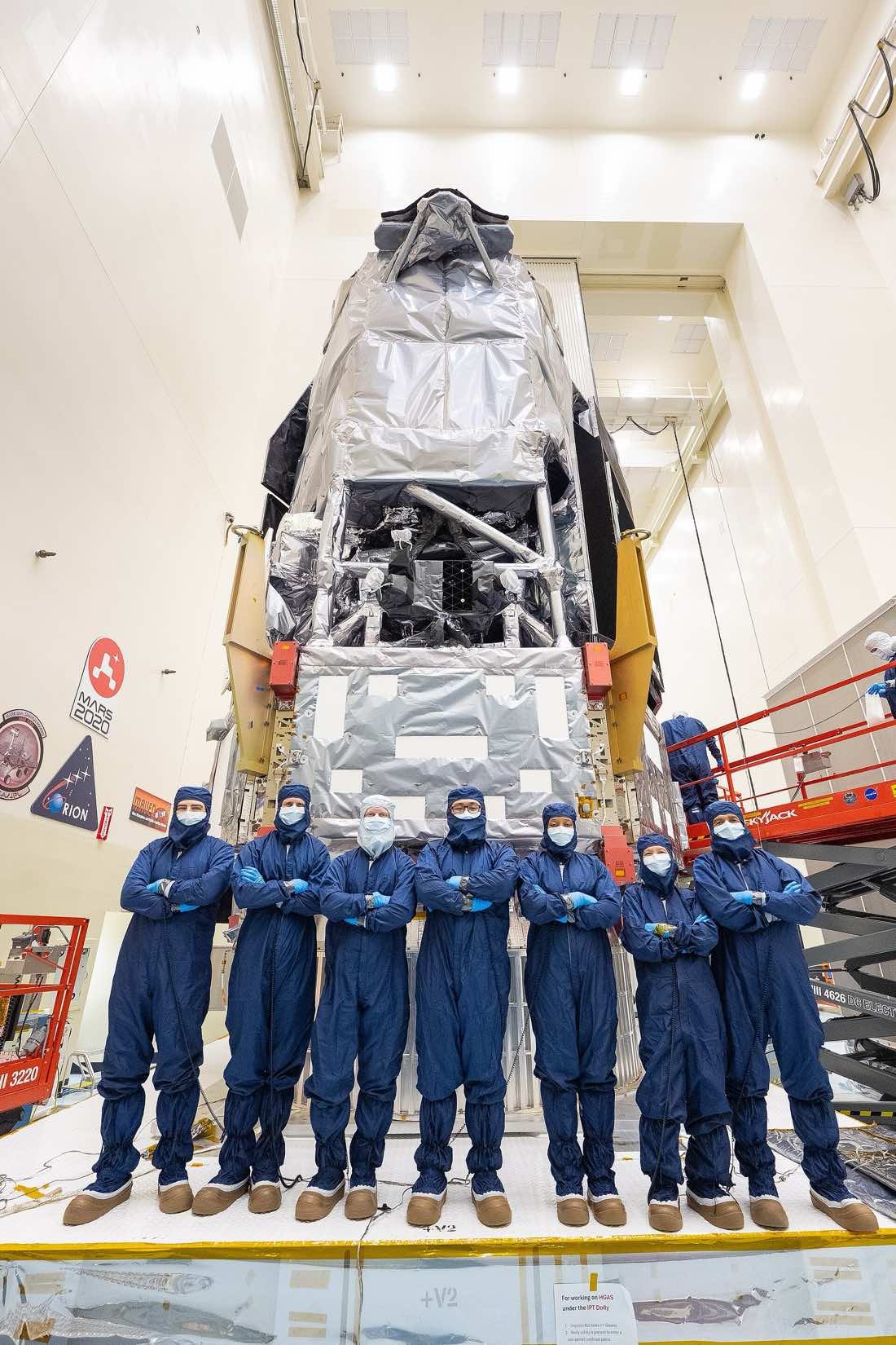 Sete engenheiros em macacões azuis de sala limpa, com touca, máscara e luvas, posam de braços cruzados diante do telescópio espacial Roman, uma estrutura alta envolvida em mantas térmicas prateadas, dentro de uma sala limpa do Centro Espacial Kennedy.