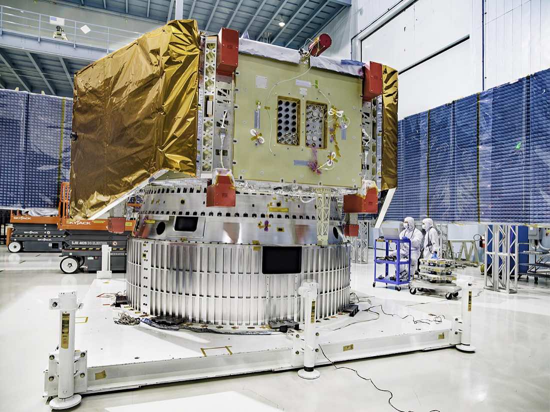 Módulo de serviço do telescópio Roman montado sobre suportes numa sala limpa, com manta térmica dourada e dois técnicos de macacão branco ao fundo