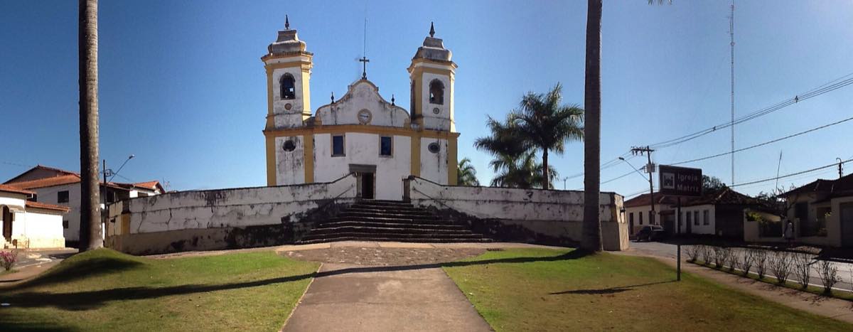 Vista panorâmica da praça e da igreja matriz de São Brás do Suaçuí, em Minas Gerais, com casario baixo ao redor e morros ao fundo.