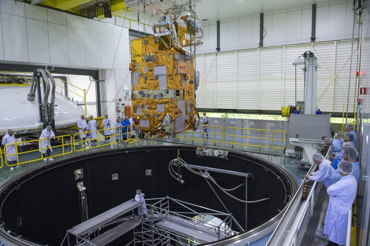 Satélite revestido de manta térmica dourada sendo baixado para dentro de uma câmara de testes, cercado por engenheiros de jaleco branco