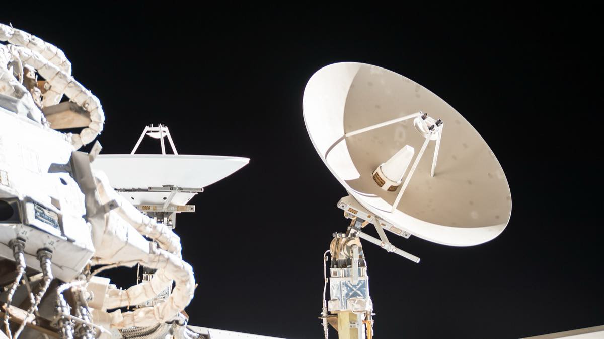 Duas antenas brancas montadas na estrutura externa da Estação Espacial Internacional contra o preto do espaço: à direita, uma parabólica grande com o alimentador sustentado por hastes; à esquerda, um refletor plano preso a um suporte junto a mantas térmicas do módulo.