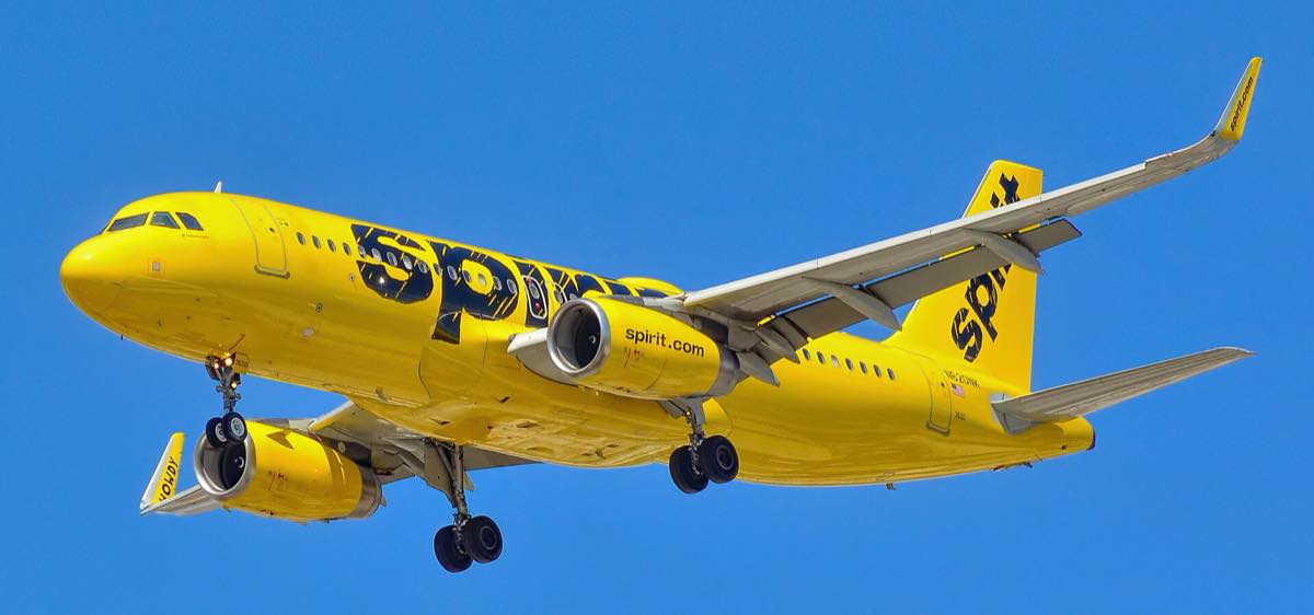 Airbus A320 pintado de amarelo com a marca da Spirit em letras pretas, em voo de aproximação com o trem de pouso baixado, contra céu azul.