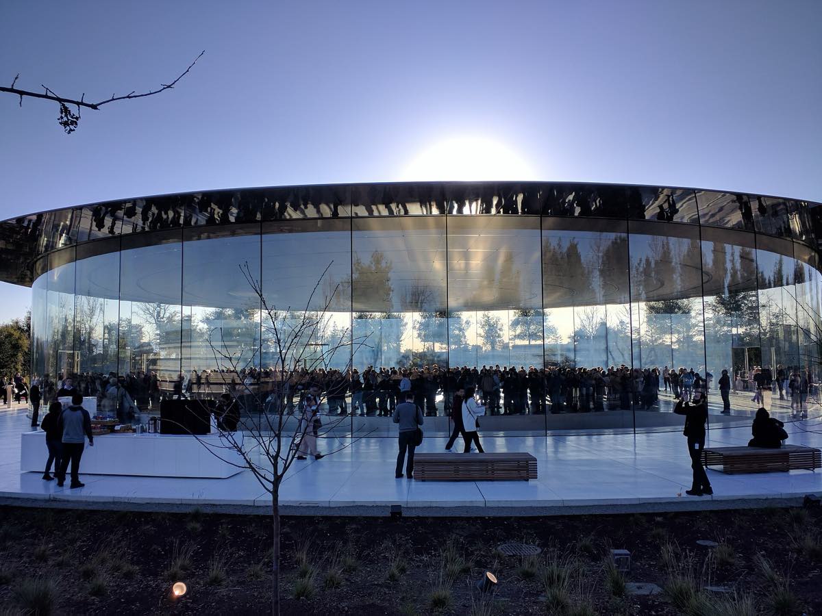 Vista externa do Steve Jobs Theater, auditório circular com paredes de vidro e teto branco em disco, no campus do Apple Park, em Cupertino