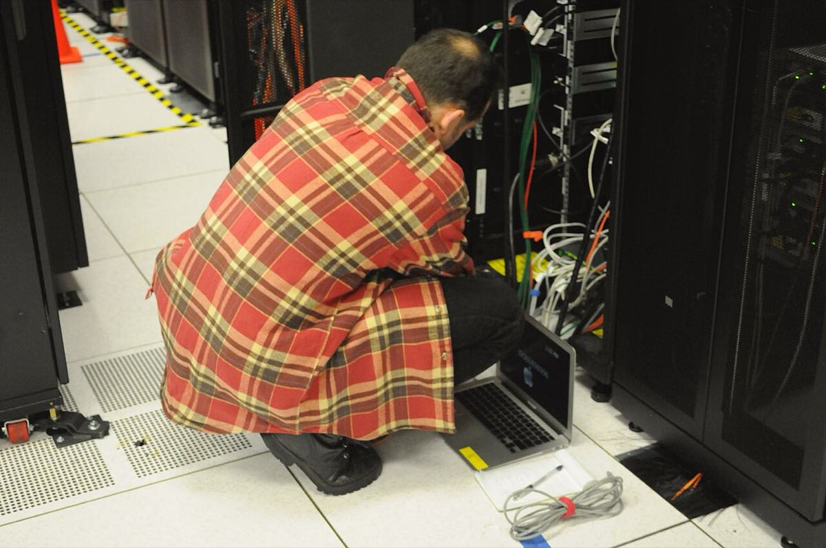 Técnico agachado no corredor de um data center, com um notebook aberto no chão, conectado a um rack de servidores cheio de cabos