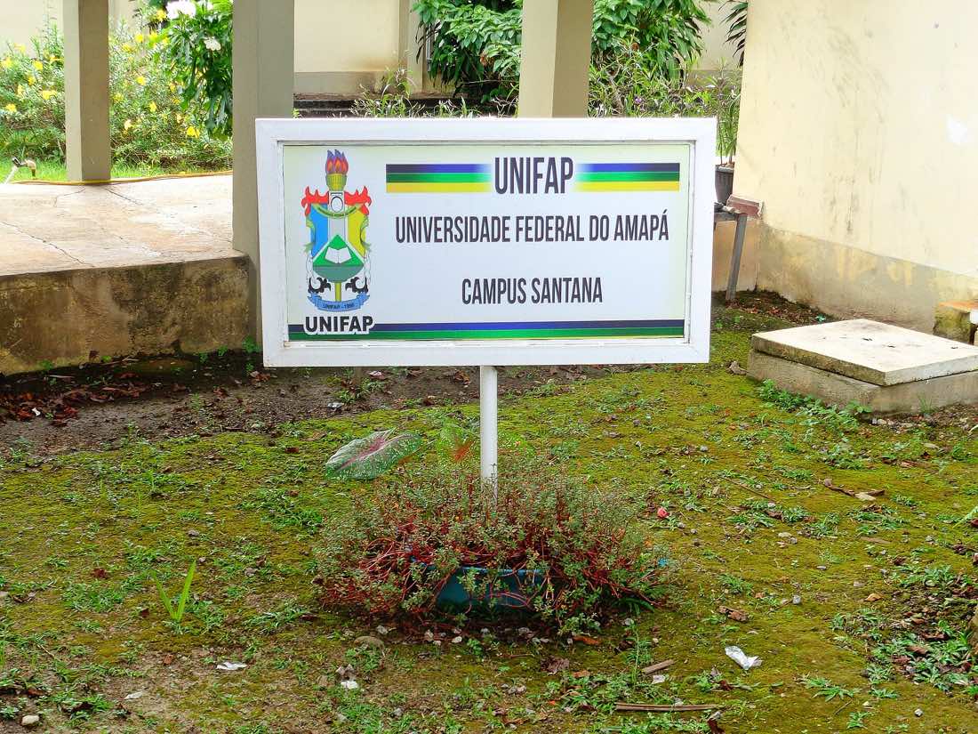 Placa branca fincada num gramado com o brasão e o nome da Universidade Federal do Amapá e a inscrição Campus Santana, diante de um prédio claro com colunas.